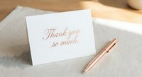 Elegant thank you card with rose gold calligraphy on white paper beside matching pen on neutral fabric background for gratitude expressions.