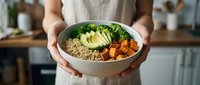 Person with medium skin tone wearing a beige apron holding a bowl with quinoa, roasted sweet potato, and fresh broccoli for healthy meal prep and nutrition.