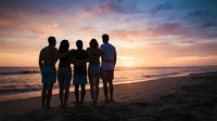 Silhouetted family of five standing together on sandy beach at sunset, watching golden sky and ocean waves, representing togetherness and vacation moments.