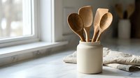 Wooden kitchen utensils in ceramic holder on marble countertop near window. Natural light illuminates cooking spoons for culinary preparation and home decor.