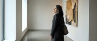 Young woman with long dark hair, South Asian ethnicity, wearing a black blazer and shoulder bag, standing in a modern art gallery viewing abstract artwork on white walls with natural light.