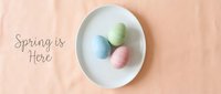 Three pastel Easter eggs on a white plate with Spring is Here text, flat lay on peach linen background for seasonal greeting design.
