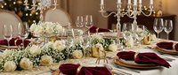 Elegant dining table set for celebration with white roses, burgundy napkins, crystal chandelier and warm lighting creating luxurious restaurant atmosphere.