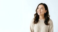 Young Asian woman with long dark wavy hair wearing a beige knit sweater, smiling and looking upward against a white background for portrait and lifestyle use.