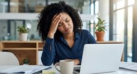 Stressed African American businesswoman with headache working on laptop in modern office, experiencing work pressure and fatigue during busy workday.