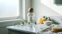 Spa wellness setup with glass bottle, natural sponge and eucalyptus on marble surface near window for relaxation and self-care routines.