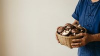 Farmer in denim shirt holding wicker basket filled with fresh mushrooms against neutral background, suitable for agricultural and food industry marketing materials.