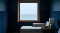 Square window with ocean view framed in wooden border on deep blue wall above white bench with lamp and laptop for interior design and home decor.