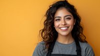 Young Latin American woman with curly hair and bright smile wearing casual grey t-shirt against vibrant orange background, expressing joy and confidence in studio portrait.