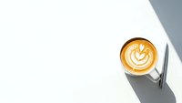 Coffee cup with latte art heart pattern on white surface with geometric shadows. Minimalist cafe scene for branding and lifestyle content.