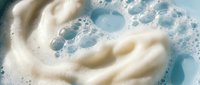 Creamy white foam swirls with bubbles on light blue background for skincare, cosmetics and beauty product advertising campaigns.