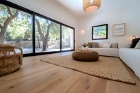 Modern minimalist living room with panoramic windows overlooking nature, featuring white sectional sofa, woven pouf, natural wood floors and neutral decor elements.