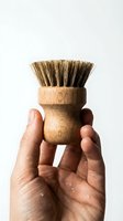 Hand holding a small round wooden dish scrub brush with natural sisal bristles against a white background, eco-friendly cleaning tool.