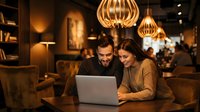Young couple working together on laptop in cozy coffee shop with warm lighting and modern pendant lamps creating intimate atmosphere for productivity.