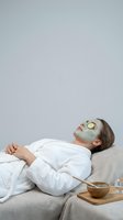 Young Caucasian woman in white robe relaxing with green clay face mask and cucumber slices on eyes during spa treatment.