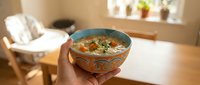 Hands holding decorative ceramic bowl with hot vegetable soup in cozy kitchen interior with natural sunlight streaming through window.