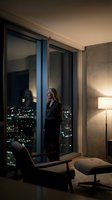 Blonde businesswoman in black suit standing by floor-to-ceiling windows overlooking illuminated city skyline at night in modern luxury apartment.