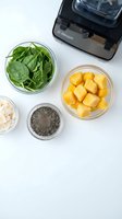 Healthy smoothie ingredients with spinach leaves, mango chunks, chia seeds and cashews arranged in glass bowls next to blender for nutritious breakfast preparation.