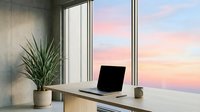 Young professional woman with medium skin tone working at minimalist desk with laptop, potted plant, and coffee cup overlooking sunset sky through floor-to-ceiling windows in modern office space.