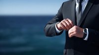 Businessman in formal suit adjusting sleeve cuff against blue ocean backdrop, portraying confidence and professional elegance.