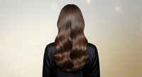 Young woman with long wavy brown hair from behind wearing black top against golden bokeh background for hair care and beauty concepts.