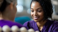 Young African American female healthcare worker in purple scrubs smiling warmly while having conversation with colleague in modern medical facility.