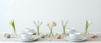 Spring Easter table setting with white dishes, pastel eggs, and fresh tulip bulbs sprouting green shoots on light wooden surface for holiday dining decor.
