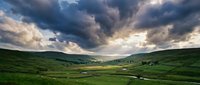 Dramatic stormy sky with golden sunlight breaking through dark clouds over rolling green hills and rural valley landscape with winding stream.