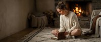 Young woman in cozy sweater sitting cross-legged on carpet near fireplace in warm living room, practicing meditation and mindfulness at home.