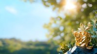 Young woman with long brown hair, wearing casual summer clothing, holding a golden trophy or award while standing outdoors in bright sunlight with blurred green foliage and hills in background.