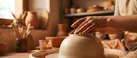 Potter hands shaping clay on spinning wheel in warm sunlit ceramic studio workshop with earthenware pottery tools and finished vessels in background.
