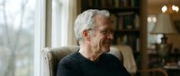 Senior man with white hair and glasses sitting by window in contemplative pose, looking outside at natural light in home interior setting.