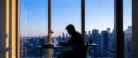 Silhouette of businessman working on laptop in modern office with panoramic city skyline view during sunset. Corporate professional lifestyle concept.