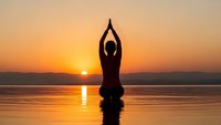 Woman practicing yoga meditation pose at sunset by calm lake with golden sky reflection creating peaceful wellness moment for mindfulness and spiritual balance.