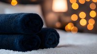 Soft navy blue rolled towels on white surface with warm bokeh lights in background creating cozy spa atmosphere. Perfect for wellness and relaxation concepts.