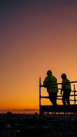 Construction workers in safety gear silhouetted against dramatic sunset sky on scaffolding platform overlooking city skyline for industrial projects.