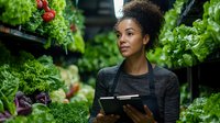 Young African American woman with natural curly hair shopping for fresh vegetables in grocery store produce section with tablet for healthy lifestyle.