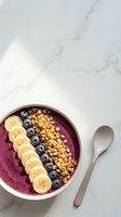 Acai bowl with banana slices, blueberries, and granola in white ceramic bowl on marble surface, ready for breakfast or healthy eating content.