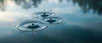 Water droplets creating ripples on calm lake surface with reflection of trees and sky in serene natural environment for meditation and wellness concepts.