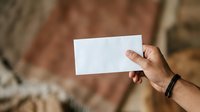 Mature male hand holding blank white card against blurred brick wall background for mockup and branding purposes.