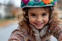 Young child wearing colorful safety helmet and patterned sweater smiles brightly during outdoor activity, showcasing joy and active lifestyle in natural lighting.