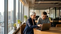 Mature woman with gray hair and younger man collaborating on laptop at wooden desk near floor-to-ceiling windows in modern office with city skyline view, representing mentorship and teamwork.