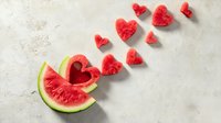 Fresh watermelon slice with heart-shaped pieces arranged in a flying pattern on light concrete background, perfect for summer food concepts.