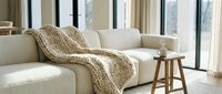 Cozy living room corner with cream upholstered sofa, knitted throw blanket, wooden side table, and natural light from large windows for home decor and interior design projects.