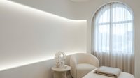 Modern minimalist interior with curved beige chair, round table and arched window. Clean white walls with LED strip lighting create serene atmosphere.