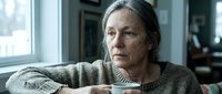 Mature woman with long gray hair and weathered facial features wearing a knit sweater, sitting indoors with contemplative expression, suitable for editorial, healthcare, aging, and lifestyle content.