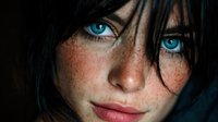 Close-up portrait of young Caucasian woman with striking blue eyes, freckled skin and dark hair covering part of face against black background.