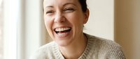 Happy middle-aged Caucasian woman laughing joyfully in cozy beige sweater at home, expressing genuine happiness and positive emotions indoors.