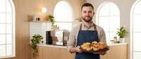 Male baker with beard holding fresh baked croissants in modern bakery shop with wooden interior, arched windows and shelves displaying products for bakery business.