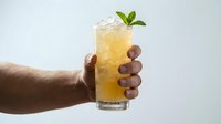 Cropped hand holding tall glass of chilled yellow beverage with ice cubes and fresh mint leaf garnish against white background, suitable for drink advertising and hospitality marketing.